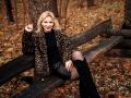 NatalyFoxy69 with blonde hair, leopard coat, black turtleneck, leather skirt, black knee boots, sitting on wooden bench, smiling, autumn forest, holding coat, showing her ass.