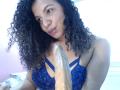 TiaraKitty with curly black hair, blue lace bra, holding a large, erect, uncircumcised dildo, smiling gently.