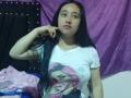 KaticaSole with long black hair, pink lipstick, white graphic tee, holding phone, showing tattoo on arm, purple and black background, pink blanket on left.