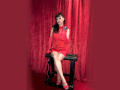 MaitresseElsa with pale skin, black hair, and bangs, wearing a red, tight, sleeveless dress, and red gloves, sits on a black bench, holding a leash, legs crossed.