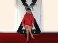 MaitresseElsa, with red mesh dress, sits on black X-shaped chair, legs crossed, holding dress hem, wearing red high heels, on red carpet. She has short black hair and red lipstick.
