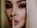 Anjellina's face, full of makeup, with blonde hair, brown eyes, and pink lips, sucking on her orange-painted finger, showing off her pouty ass.
