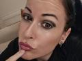 GennyRock with dark eyes, heavy eyeliner, and pink lipstick, biting her finger with a gold lip piercing, black shirt, and ear studs, looking at the camera.
