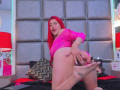 AlissaBrown with red hair, wearing a pink sheer top, is on a bed, holding a black dildo in her ass, her tongue out, looking sexy.