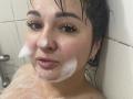 AlinaMilf's wet face with soap suds on her cheeks and chin, dark wet hair, brown eyes, and slightly parted pink lips in a bathroom. Her skin is fair and she's wearing gold earrings.