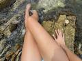 SandraSwweet's legs crossed, wet, over rocky water, hands with pink nails resting on her thighs, sandy skin, toes in water, small rock at her side, water rippling.