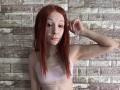 HannaCool with red hair, freckles, pink sports bra, and a nose ring, raising her right arm, showing her bare tits, against a textured brick wall.