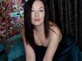 MarleneDavies with pale skin, blue eyes, and dark brown hair, leaning forward in a black strapless dress, showing her tits, in a dimly lit room.
