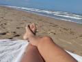 LaFrancaise's legs with tan skin, crossed at the ankles, lying on a white towel on a sandy beach, feet pointing towards the ocean with small waves.