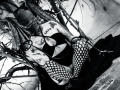 AmySpider with tattoos, wearing fishnet stockings and black top, squatting between tree branches, black-and-white, showing her tits and arms, wearing glasses, with dark makeup, looking at camera.