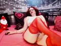 TereseHot with red lace stockings, red lipstick, and glasses, lying on a pink bed, exposing her tits and pussy, legs raised, Santa toy on left, black pillows with hearts.