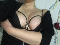 LadyMiller with large, round boobs in a black bra with strappy details, arms crossed, wearing a black cardigan, light skin, no face visible, necklace, red heart decoration.