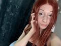 HannaCool with red hair, freckles, nose ring, lip piercings, and eyeliner, touching her hair, wearing a black top, showing her tits, in a selfie.