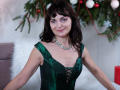 AngieGreen has green satin dress with lace-up front, showing her tits, wearing a sparkly necklace, smiling with pink eyeshadow, brown wavy hair, and white furry pillow in background.