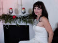 AngieGreen with dark brown hair, blue eyes, and pink lipstick, wearing a white strapless dress with a sparkly necklace, sitting in front of a decorated fireplace.