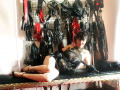 MaitresseElsa lying on a black bench, wearing a sheer black dress with gold embroidery, black high heels, and black gloves, surrounded by various black and red BDSM gear.