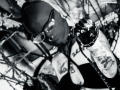 Black-and-white photo of AmySpider with a bald head, glasses, ball gag, and leather harness, arms bound with ropes, tattoos visible on arms, lying on a tangled surface.