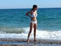 LaFrancaise stands on a pebble beach, wearing a white bikini with her hands on her hips, showing her tits and ass, looking at the ocean.