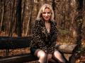 NatalyFoxy69, blonde, sitting on a wooden bench, wearing a leopard print jacket, black skirt, and sheer pantyhose, with her hands on her thighs, in a forest.