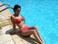 JoliDanielle with wet brown hair, wearing a red lace bikini, sitting by a blue pool, her ass and small tits visible, green wristband, legs in water, looking down.