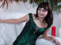 AngieGreen with dark brown hair, green satin corset showing tits, lace-up front, silver necklace, white fur, red candle, and Christmas decor, leaning on white couch, smiling.