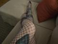 GennyRock's legs in black fishnet stockings, knees up on a beige couch, showing a tattoo on her thigh, with a red pillow to the right.