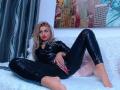 LusyBella with blonde hair, red lipstick, and black latex pants, sitting on a white fluffy couch, legs spread, showing her shiny black pants and pink nails.