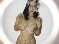 SquirtinGamy with large tits in a white, barely-there bra and matching thong, wearing a flower headpiece, has hands up, dark wavy hair, and a soft smile.