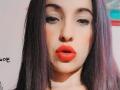 LuluFrancaise has pale skin, black straight hair with purple highlights, dark eyebrows, and bright red lipstick, with slightly parted pinkish-red lips, looking straight into the camera.